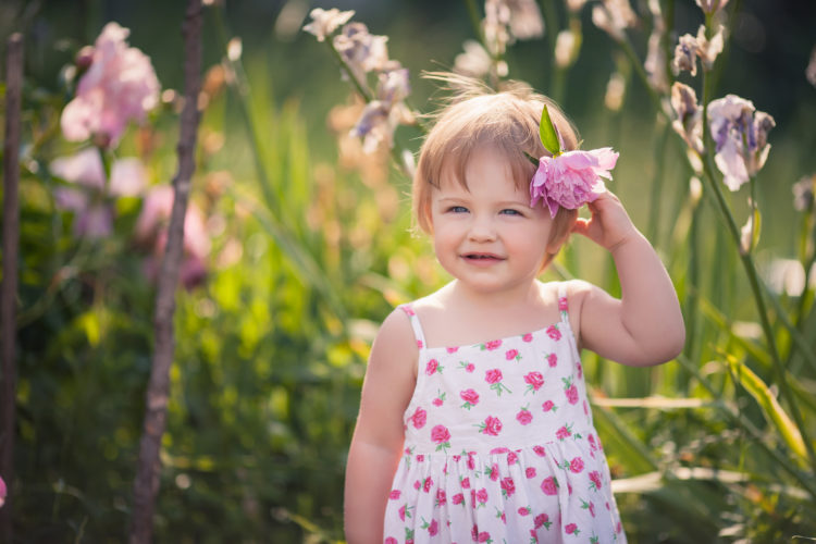 Image of baby girl with blue eyes blond hair and pink peony in her hair in summer garden with bright flowers. She dressed on white with floral pattern summer dress