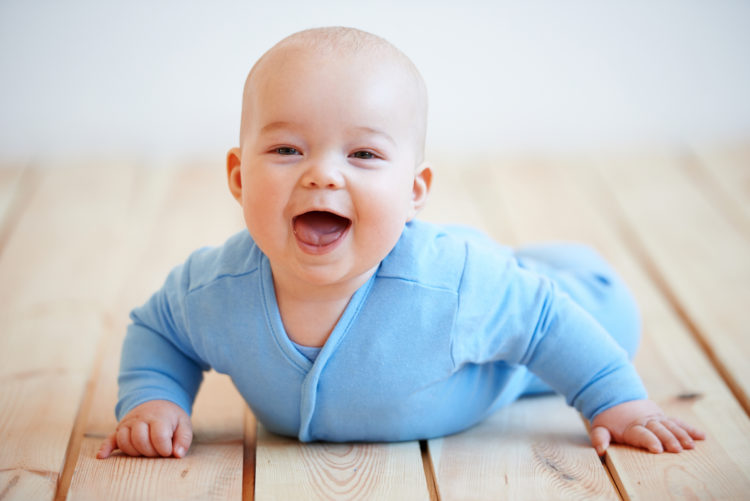 Image of Cute happy baby boy crawling on the wooden deck or floor lifting itself up on its hands to laugh at the camera