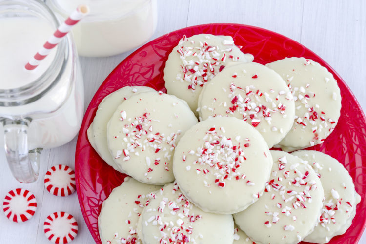 Image of Fresh baked chocolate peppermint cookies coated with white chocolate and candy cane sprinkles stacked on a festive red plate with 2 glasses of milk